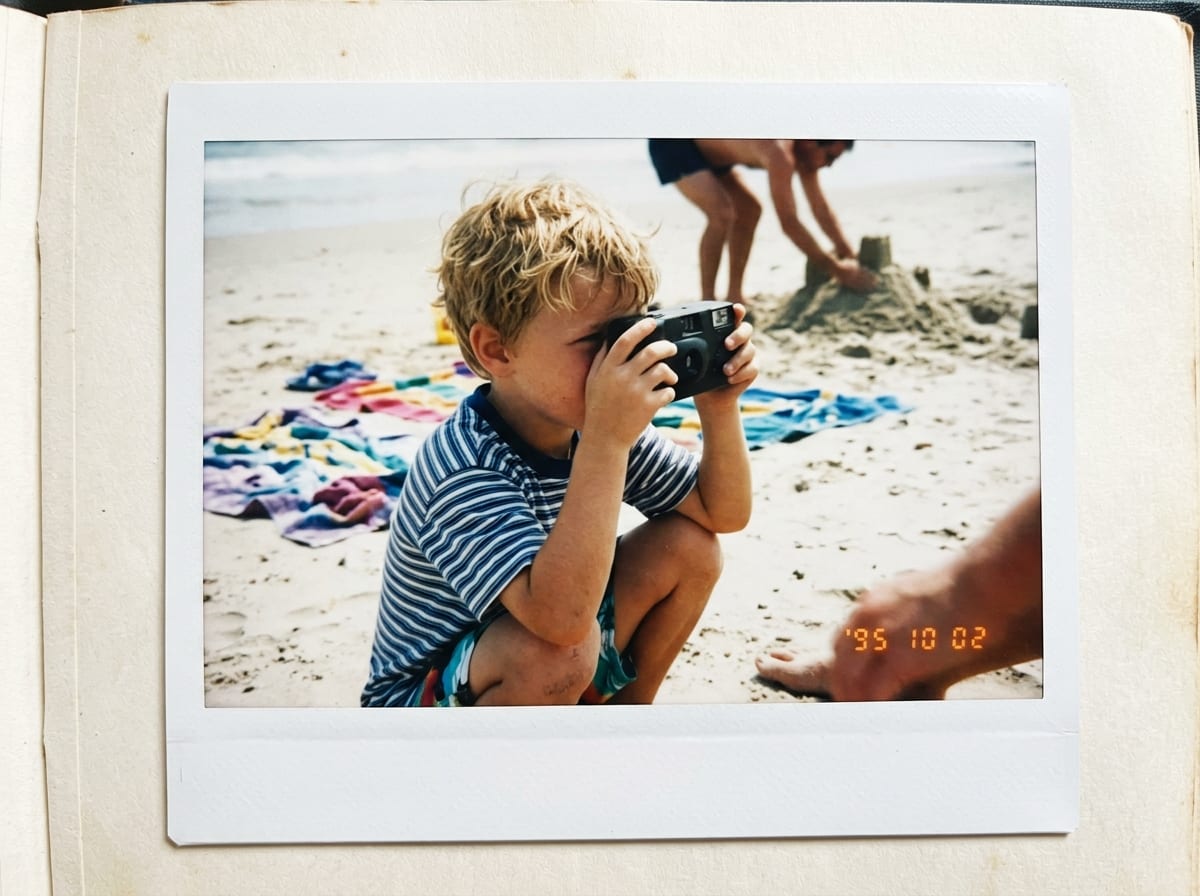 Sur une plage, un enfant photographie avec un appareil jetable son parent qui construit un château de sable.
