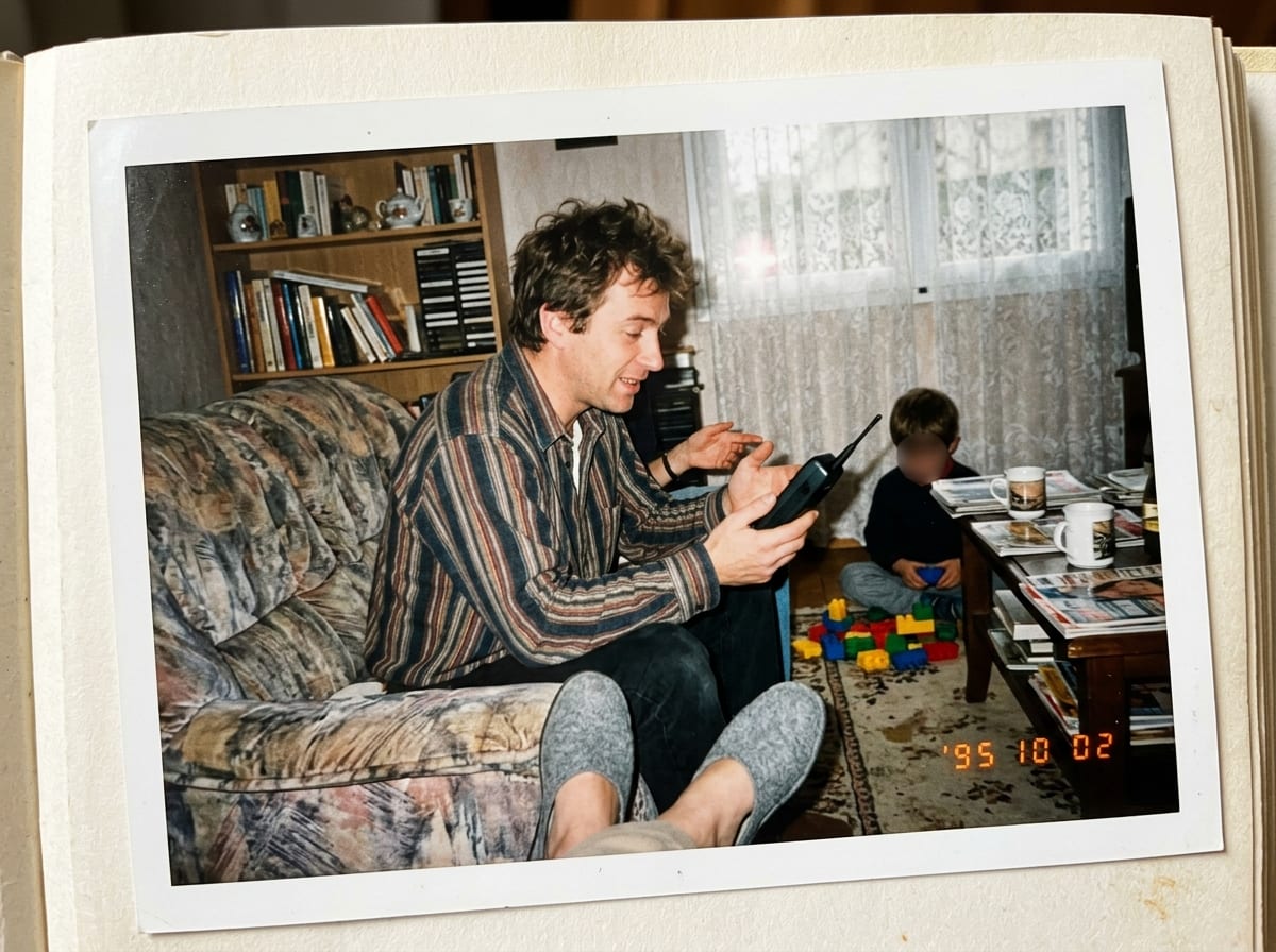 Un père assis dans le canapé du salon tient un gros téléphone portable des années 90 pendant qu'un enfant joue aux briques sur le tapis.